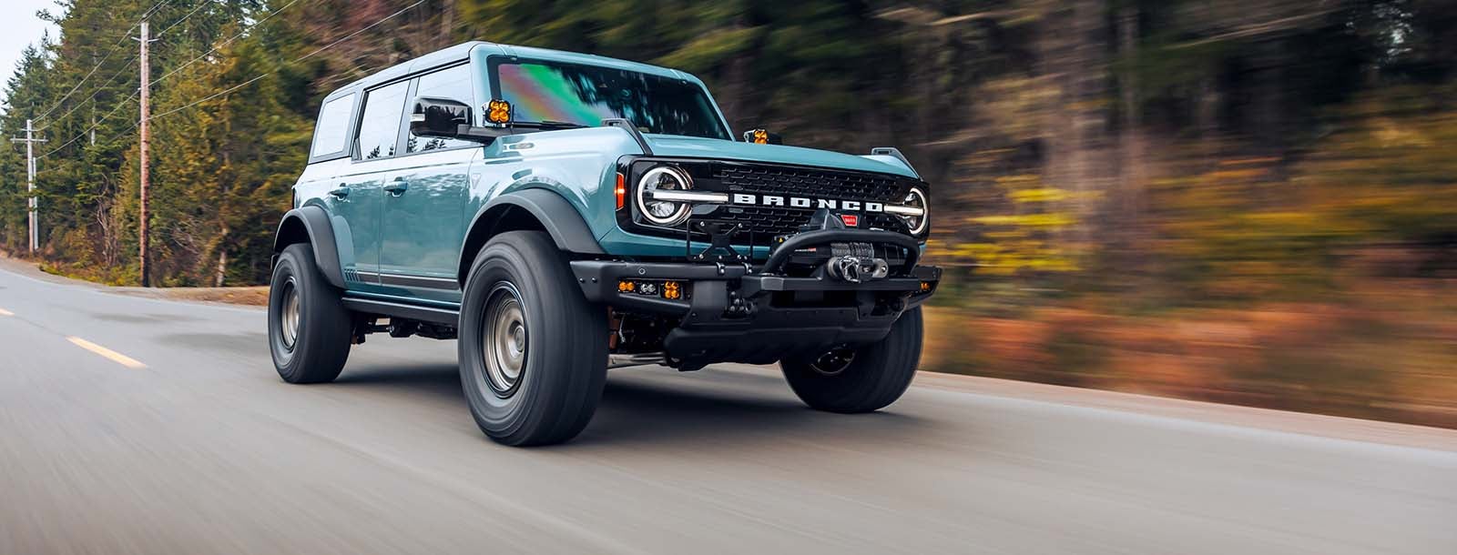 A Ford Bronco SUV driving on a scenic road with mountains in the background, showcasing its rugged and adventurous spirit.