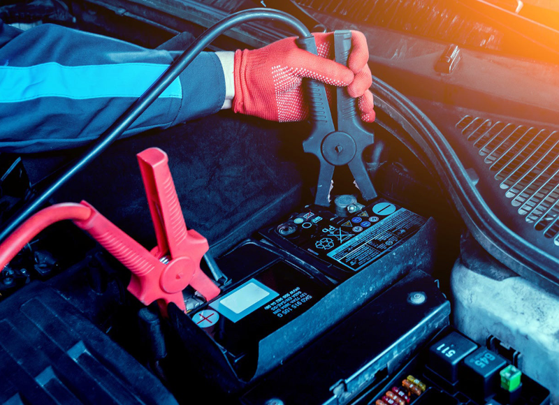 A person in red gloves attaches jumper cables to a car battery to jump-start the engine.