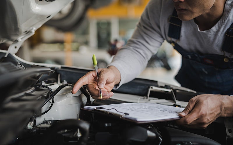 A mechanic in overalls inspects a car engine with a pen and checklist.